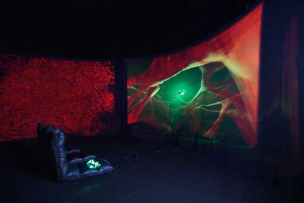 Im dunklen Inneren der Rauminstallation, gepolsterte Sitze von der Seite, leuchtende Gamekonsole und Animation am Screen